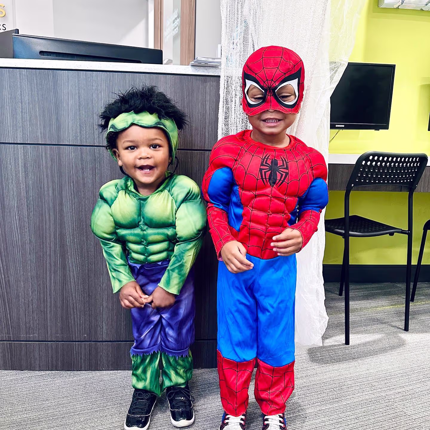 Two young children smiling and standing indoors wearing Hulk and Spider-Man costumes.
