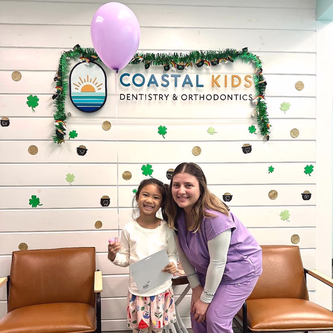 Smiling young girl holding a certificate and pink balloon, standing next to a woman in purple scrubs in a dental office decorated with shamrocks and gold coins.