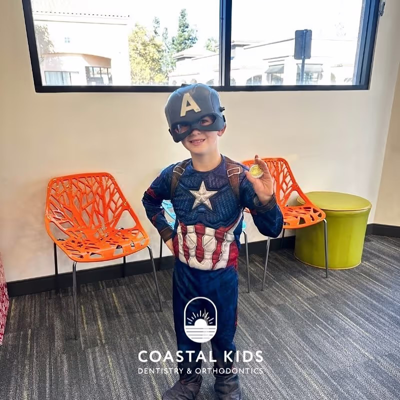 Young child dressed in a Captain America costume inside a dental office holding a gold coin and smiling.