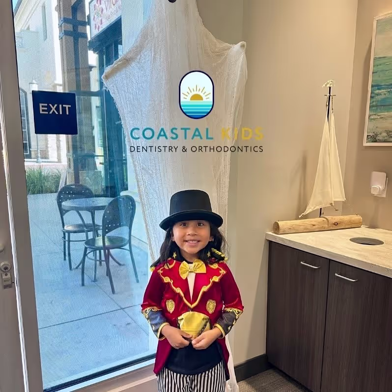 Smiling child dressed as a circus ringmaster with a black top hat, red jacket with gold accents, standing inside an office near a glass door with an exit sign.