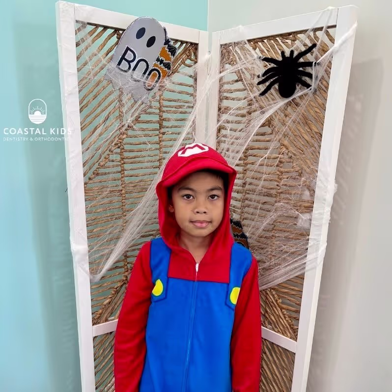 Child dressed in a red and blue Mario costume standing in front of a Halloween-themed screen decorated with a ghost and spider web.