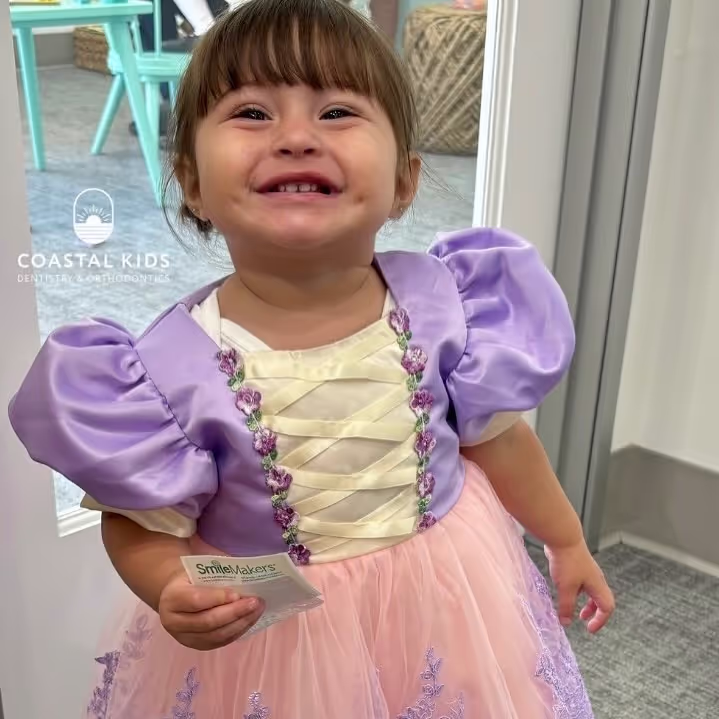 Smiling toddler girl wearing a purple and pink princess dress holding a small packet, standing indoors near a door.