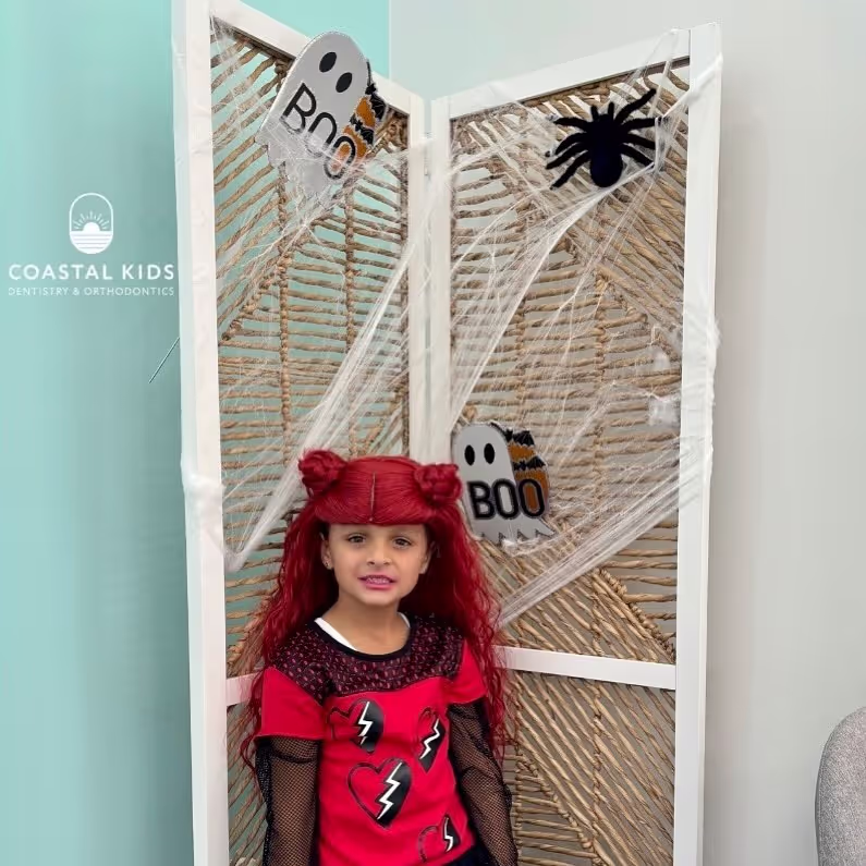Young girl with red hair in Halloween costume standing in front of a decorative screen with spider webs, ghosts, and a spider.