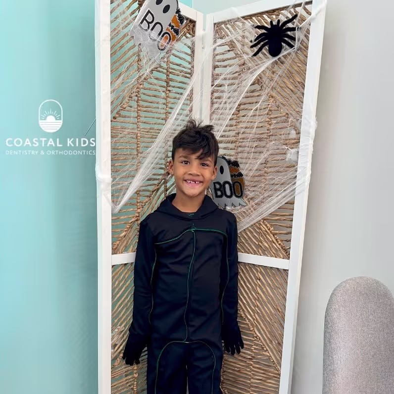 Smiling boy in black Halloween costume standing in front of a decorative screen with fake spider webs, a black spider, and cutouts that say 'Boo'.