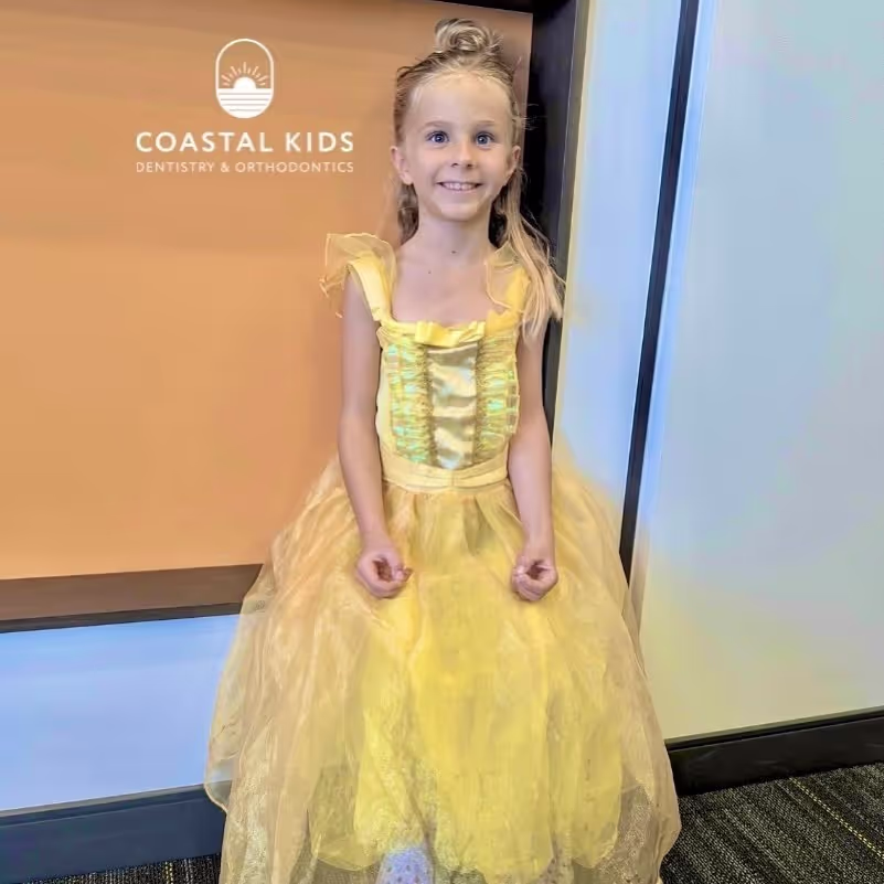 Smiling young girl in a yellow princess dress standing indoors against a wall with Coastal Kids Dentistry & Orthodontics logo.