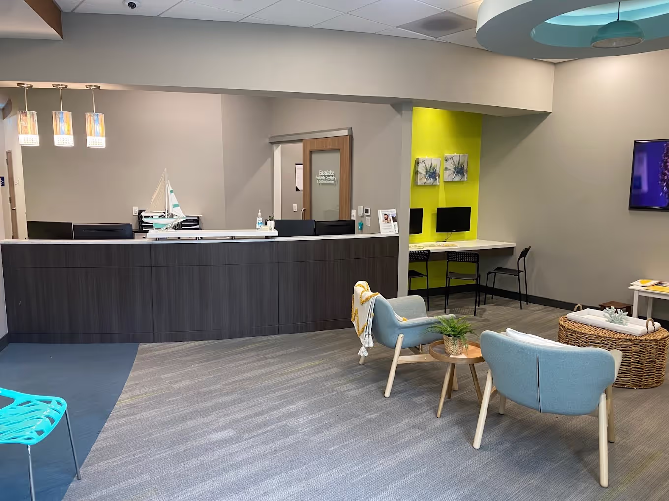 Modern office reception area with a dark wood front desk, two light blue chairs, a small table with a plant, a computer workstation against a bright yellow accent wall, and a TV mounted on the right wall.