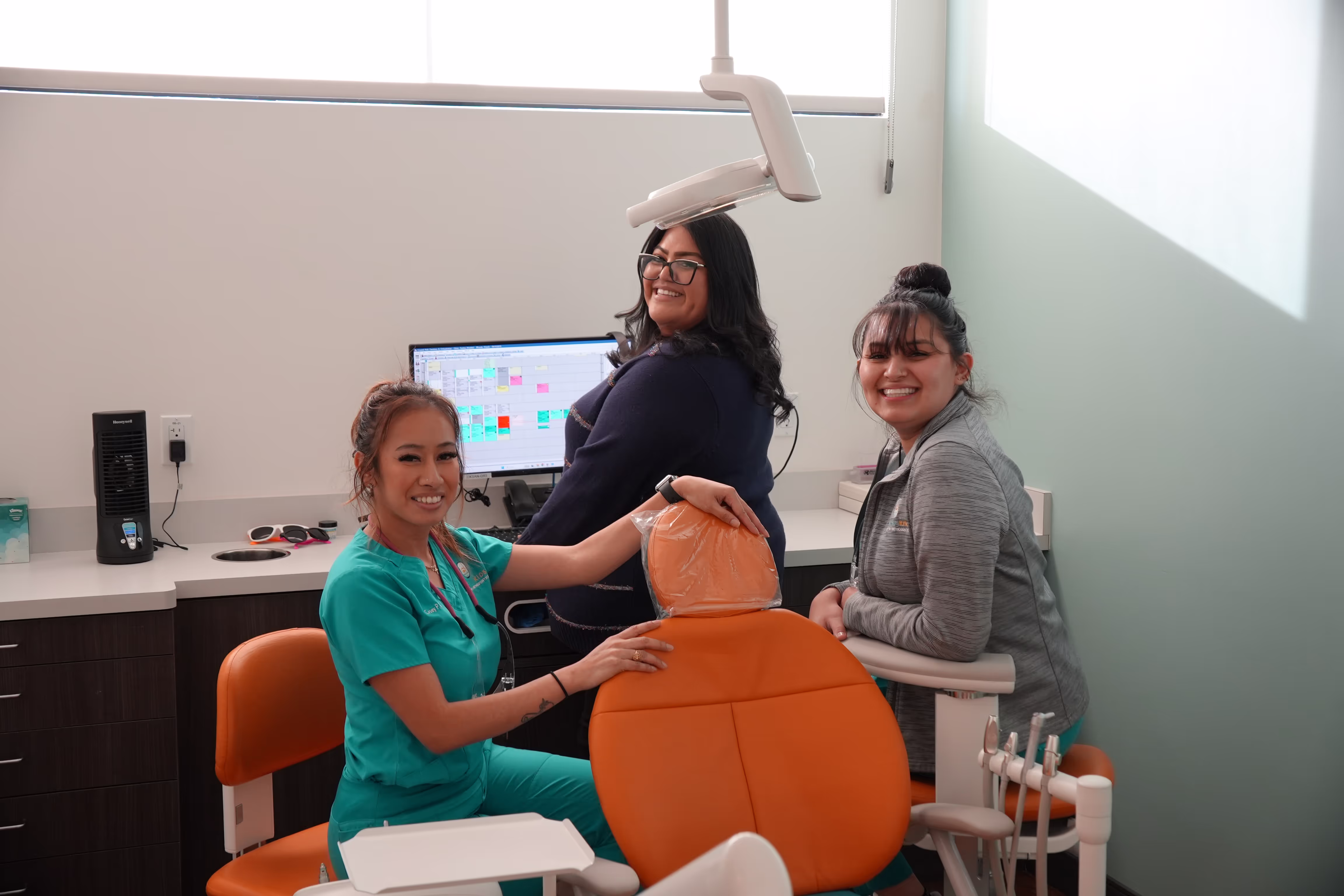 Three smiling women in a dental office, one in teal scrubs seated by an orange dental chair, others standing nearby with a computer and dental equipment visible.