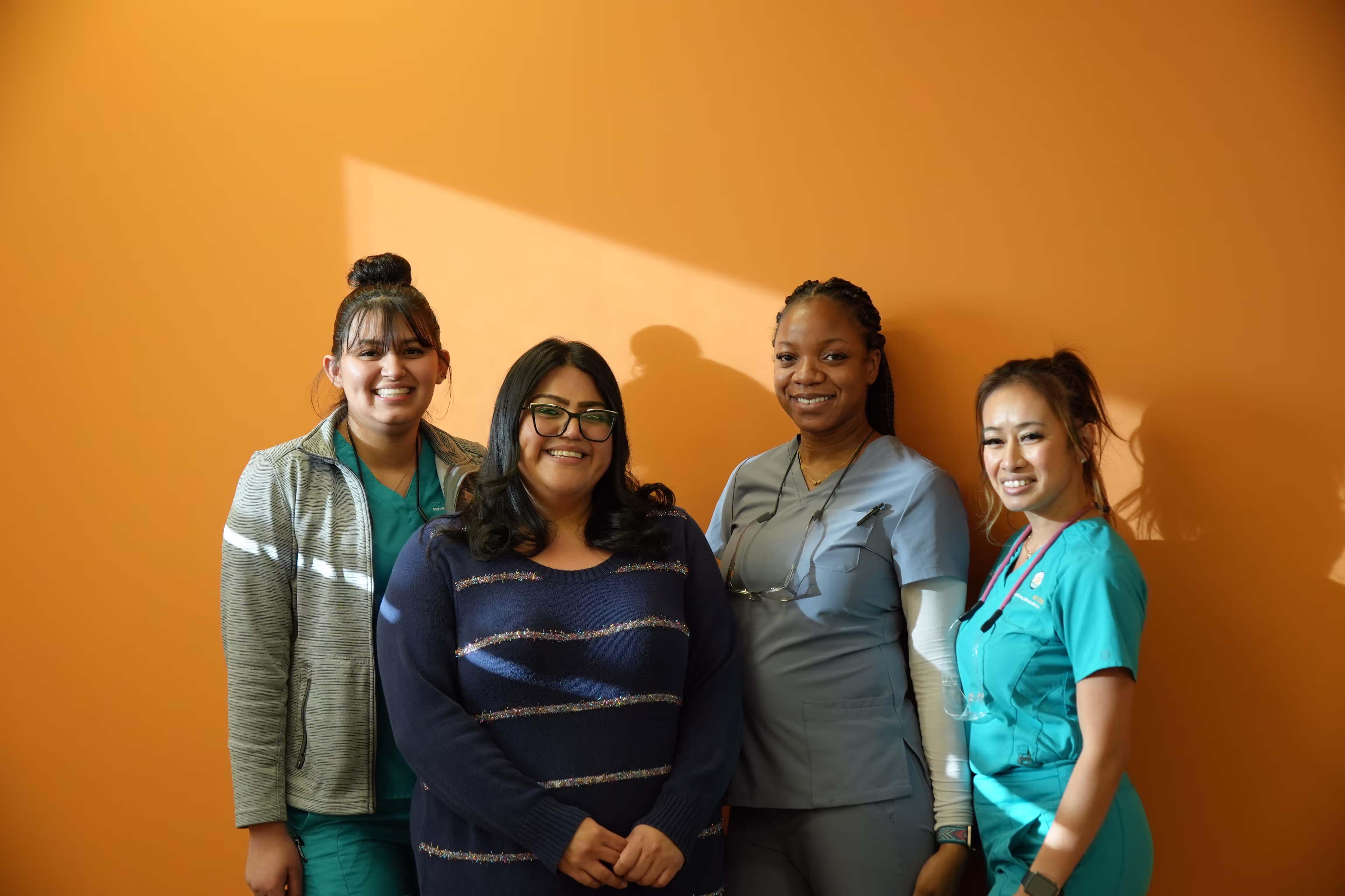 Four smiling women standing in front of an orange wall, three wearing medical scrubs and one in a navy blue sweater.