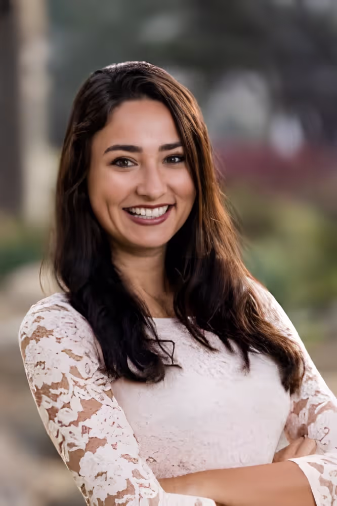 Smiling woman with long dark hair wearing a white lace top, standing outdoors with arms crossed.