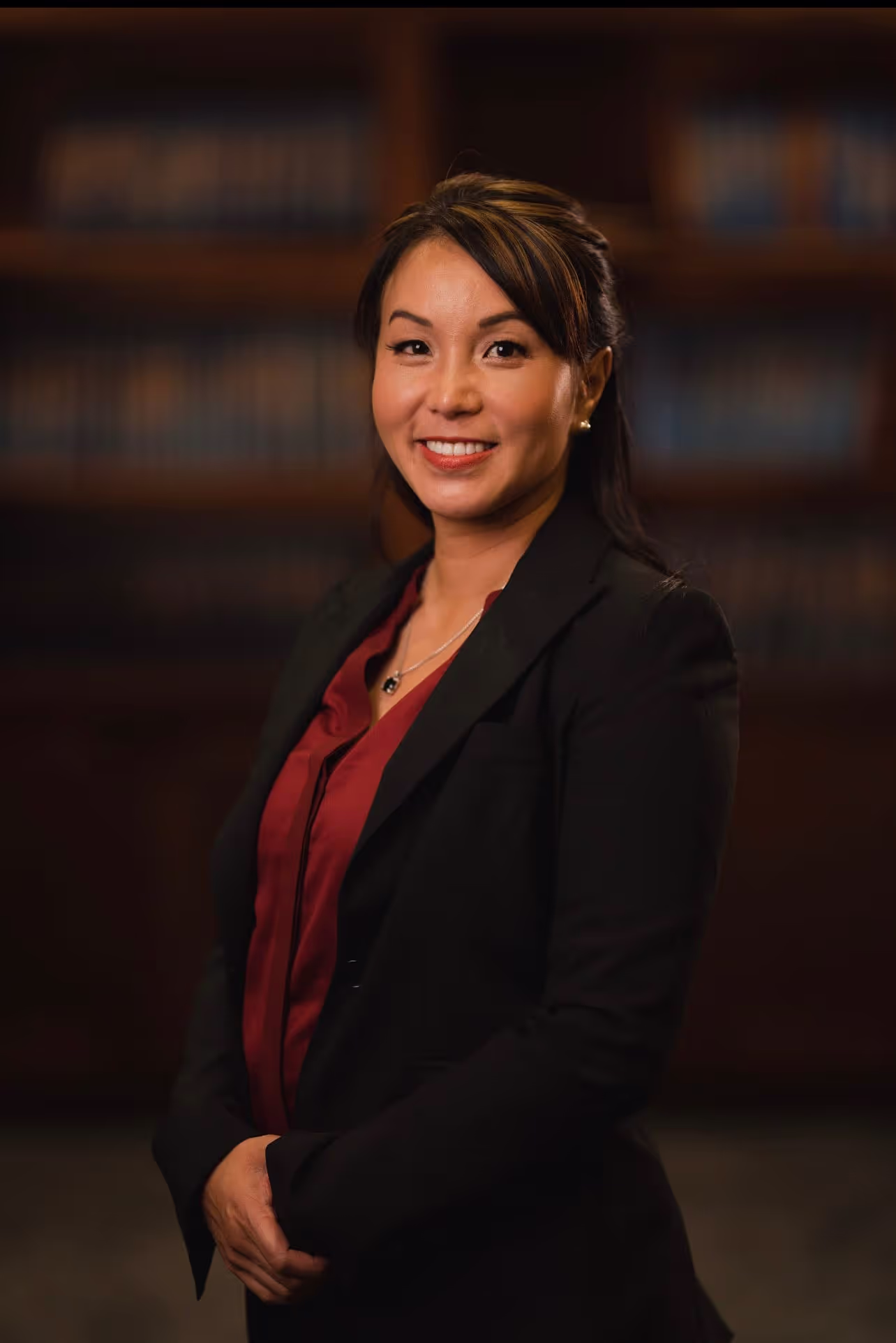 Smiling Asian woman wearing a black blazer and maroon blouse standing in front of a blurred bookshelf background.