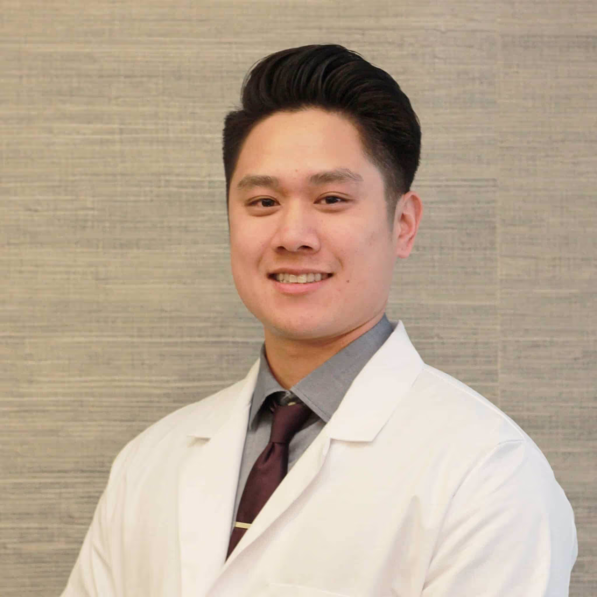Smiling man wearing a white lab coat, gray shirt, and dark tie standing against a textured beige background.
