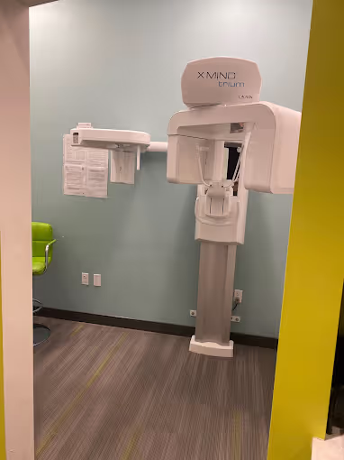 Dental X-ray machine in a clean, small room with light blue walls and a green chair in the corner.
