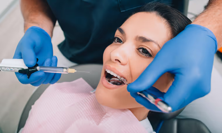 Dentist wearing blue gloves preparing to administer an injection to a smiling woman receiving dental treatment.