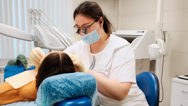 Dentist wearing mask and magnifying loupes examining a patient reclined in a dental chair.