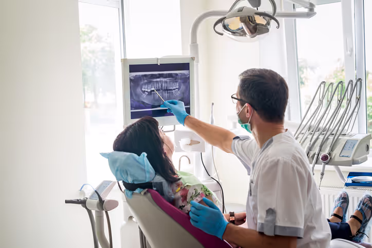 Dentist wearing gloves and a mask showing a dental X-ray on a screen to a female patient seated in a dental chair.
