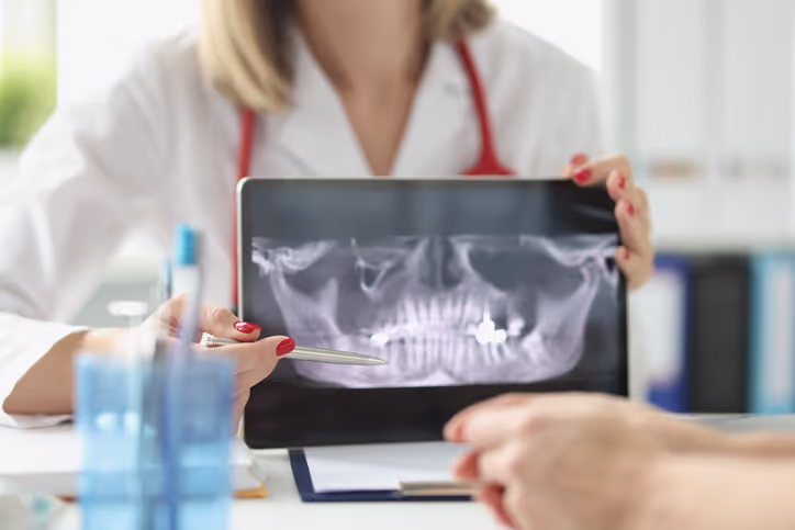 Dentist showing a dental X-ray on a tablet to a patient and pointing with a pen.