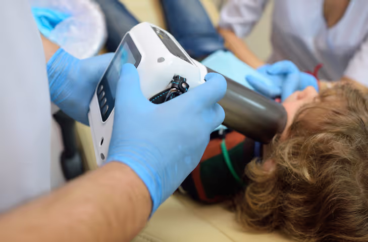 Medical professional wearing blue gloves holding a handheld X-ray device near a patient's face lying down.