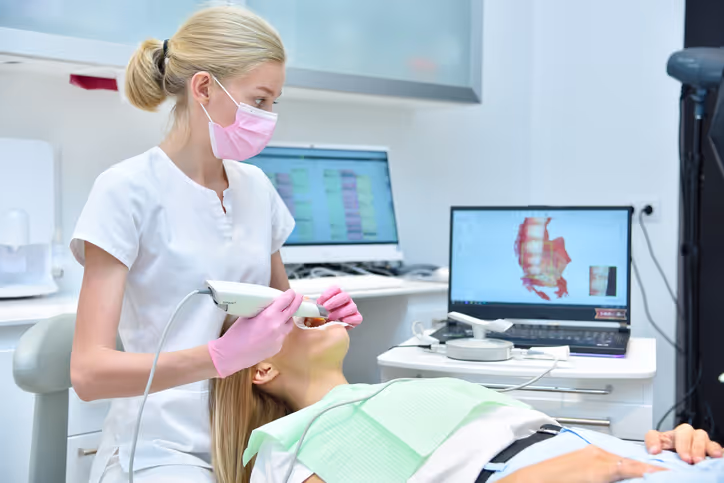Dental professional wearing a pink mask and gloves using an intraoral scanner on a patient lying in a dental chair with 3D dental images displayed on a laptop screen.