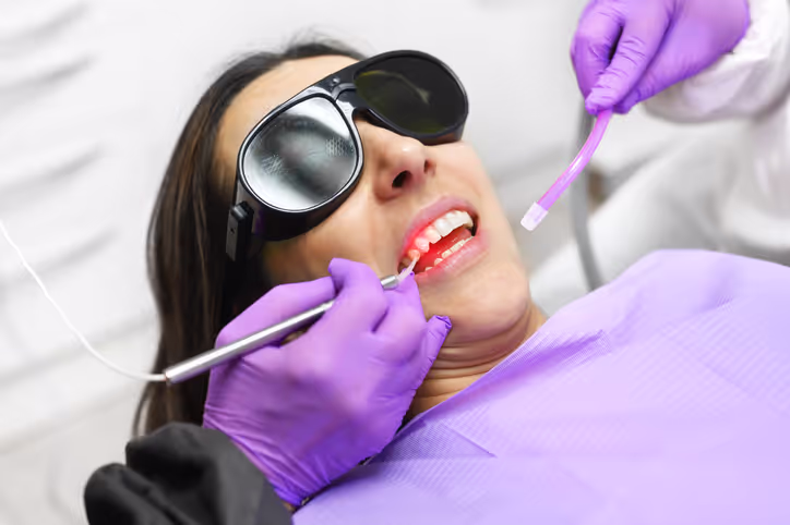 Patient wearing protective glasses receiving dental laser treatment from dentist with purple gloves.