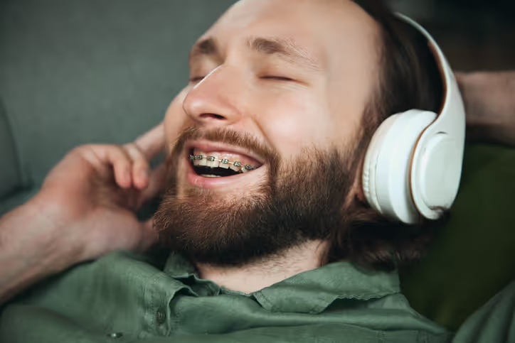 Bearded man with braces smiling and enjoying music while wearing white noise-canceling headphones.