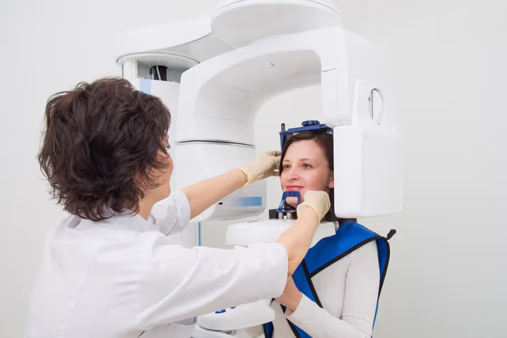 Dental technician adjusting a woman’s head positioning in a panoramic dental X-ray machine, patient wearing protective lead apron.
