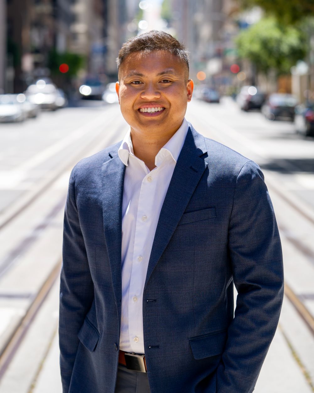 Smiling man wearing a navy blue blazer and white dress shirt standing on a sunlit city street with tram tracks.