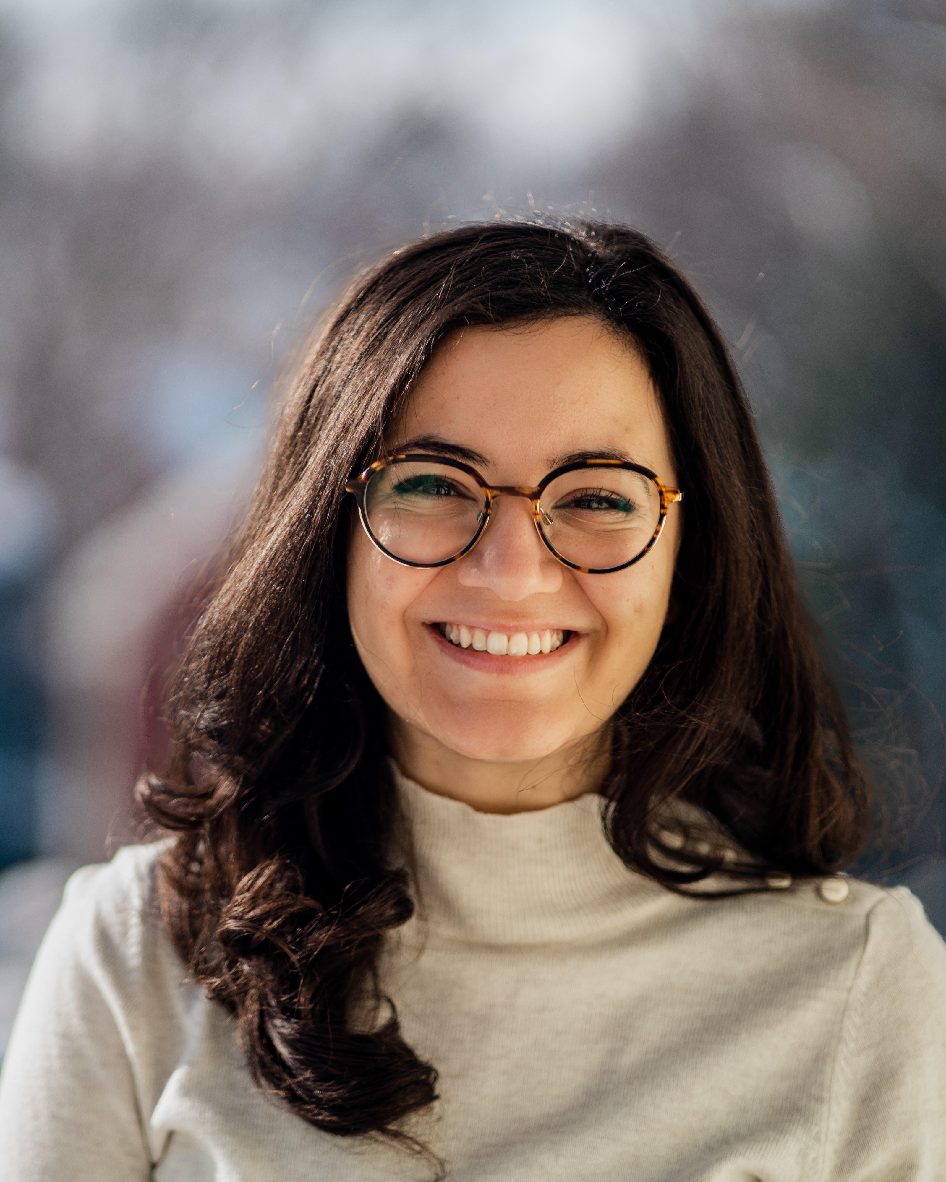 Smiling woman with long dark hair, wearing tortoise shell glasses and a light beige sweater.